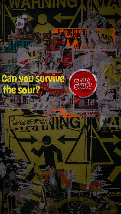 An ad for Dead Sour created by Ayanna Richilano Designs with the slogan 'Can you survive the sour?' and logo type plastered on a faded wall covered in posters  to help the design pop out from the chaos.