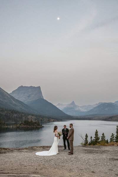 A couple stands with their officiant along the edge of a mountain lake at twilight, surrounded by soft evening light and alpine peaks in Glacier National Park, captured by Sydney Breann Photography during their intimate elopement.