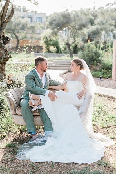 Elegant bride and groom seated on a vintage sofa under olive trees in Meganisi, Greece, during a dreamy wedding celebration