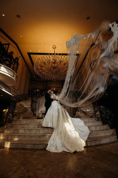 Bride and groom kiss on grand staircase as wedding veil dramatically billows behind