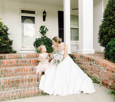 Virginia Beach wedding photography of a  bride, holding a bouquet, and a flower girl  sitting on the stairs in front of a beautiful white building and smiling at each other taken by Virginia wedding photographers Glenn and Nadya