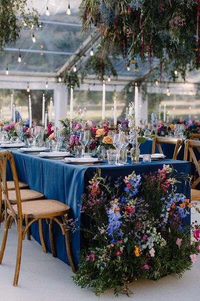 A celestial-themed wedding marquee with tables draped in deep blue, adorned with a vibrant mix of wildflowers and tall, elegant candles.
