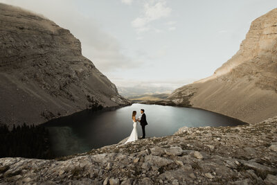 A wedding couple standing on a rocky mountain overlooking a lake.
