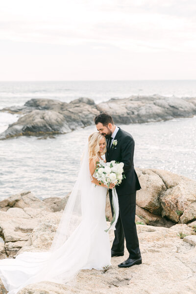 Wedding Photographer, a groom happily hugs his bride as she holds her bouquet near the ocean tides and rocks