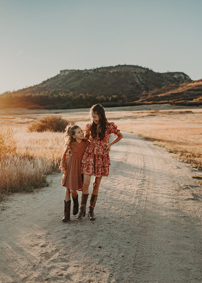 A big sister skips with her little sister while wearing rust colored dresses and cowboy boots at the base of a mountain at sunset in Colorado.