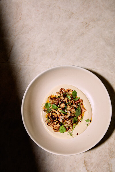 A minimalist and elegant image featuring a grilled dish garnished with fresh herbs, served in a simple white bowl. Captured by Mica McCook, this photograph emphasizes the balance between simplicity and flavor.