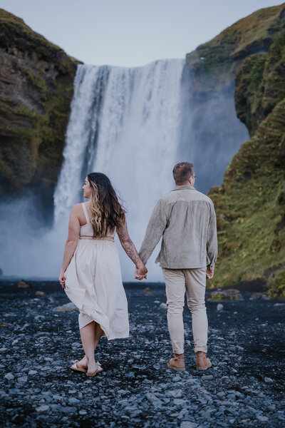 Bride and groom hold hands while walking toward Skógafoss waterfall, a dreamy Icelandic backdrop for their adventurous love.