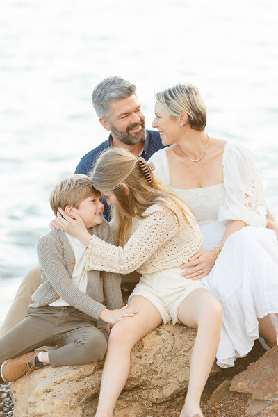 A family of 4 portrait taken at a Dallas lake while they are all sitting on a rock smiling at each other for their family photos.