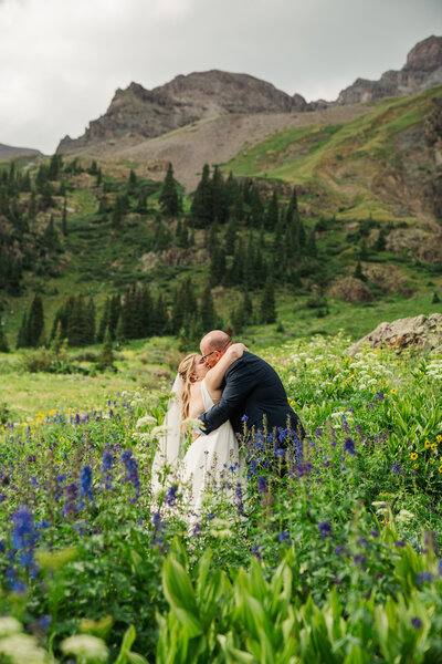 A bride and groom smile at each other while holding hands and walking towards the camera carrying skis