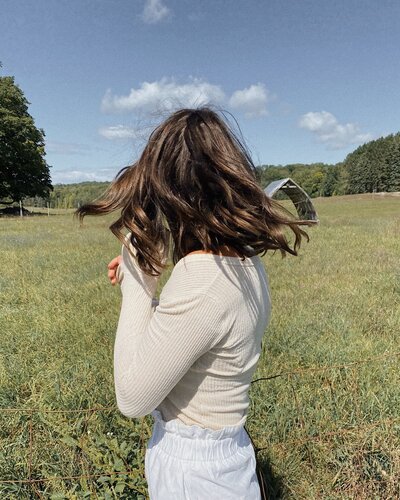 A person with shoulder-length brown hair stands in a sunny field, wearing a light long-sleeve shirt. Trees and a clear blue sky are in the background.