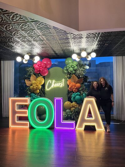 Light up neon marquee letters with two women standing to the right and a tropical themed backdrop and balloon in behind. 