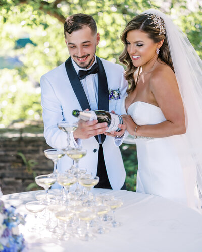 A stylish bride and groom pour champagne into a glass tower during their outdoor celebration in Pittsburgh, smiling and dressed in classic formalwear.