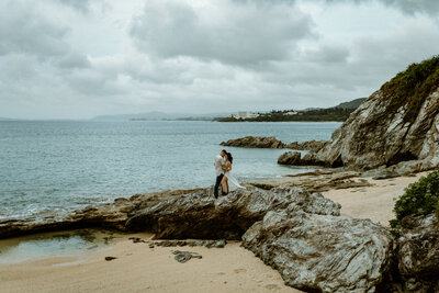 N standing behind M, arms wrapped around M with her face pressed against M's back, large landscape of them standing on a rock with the ocean behind them