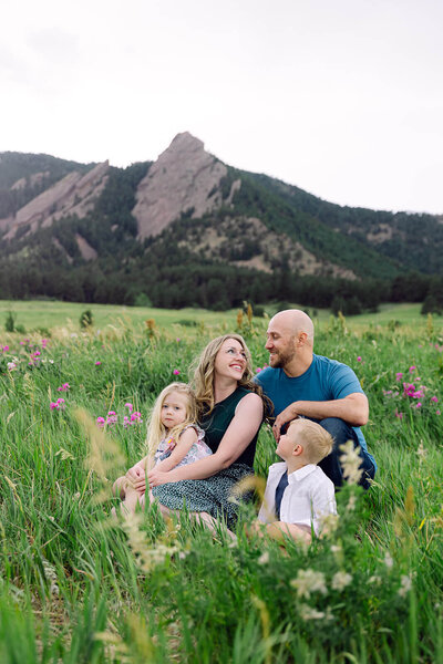 Colorful family session at Chautauqua Park in Boulder Colorado. Family of 4 smiling and looking at each other, sitting on the grass during a windy spring day.