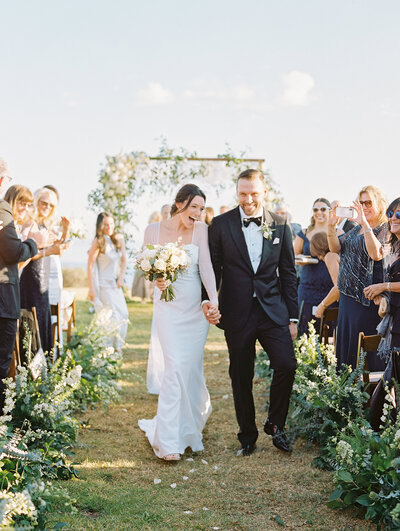 A bride and groom walk hand in hand down a grass aisle, smiling at their wedding guests as they clap. The wedding dress is body fitting, with full-length, semi-see-through sleeves.