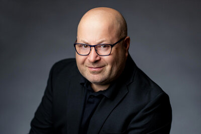 Business portrait of bald man with glasses in black shirt, confident look against dark gray backdrop