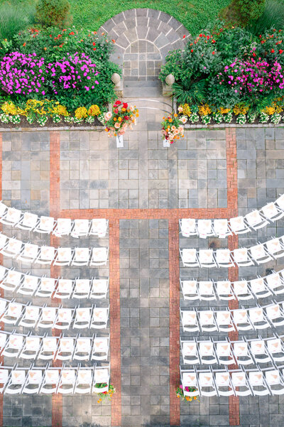 An outdoor wedding setup with rows of white chairs arranged on a patterned stone patio. Surrounding the area are colorful flowerbeds and a stone walkway leading to a small archway.