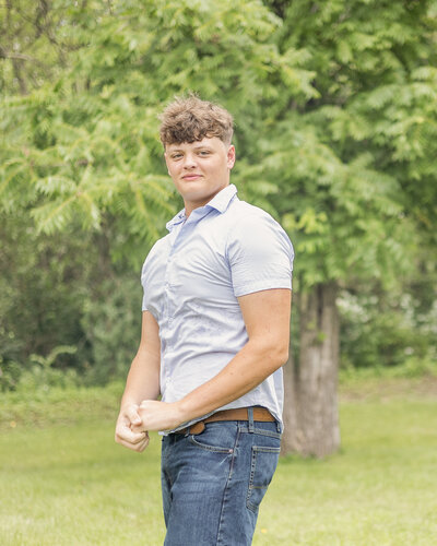 high school senior male standing in the grass, with trees in the background, turned to the side looking at the camera with a soft smile, wearing jeans and a short sleeve button up shirt