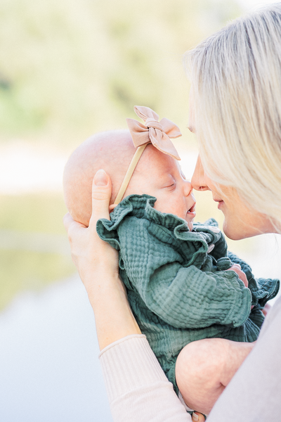 close-up photo of a newborn baby girl dressed in a dark green romper. She is held by her mother who goes nose to nose with her. A river can be seen in the background in this sweet moment captured by a photographer specializing in newborn photography in Austin, TX