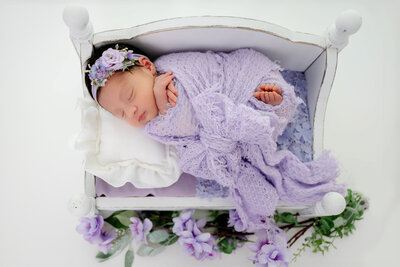 Close-up of newborn wrapped in lilac fabric tied in a bow, wearing a matching headband, lying on a white bed with flowers below.