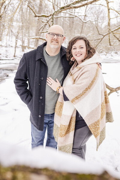 Husband and Wife hugging smiling at the camera on a Winter day out in the snow by a creek in Brown County, Ohio.