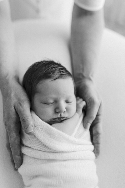 A black and white image of a newborn being held in his fathers hands during a newborn photoshoot with christchurches top newborn photographer lucy jane
