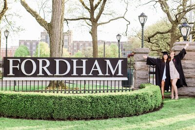 Two girls in caps and gowns posing beside a Fordham University sign