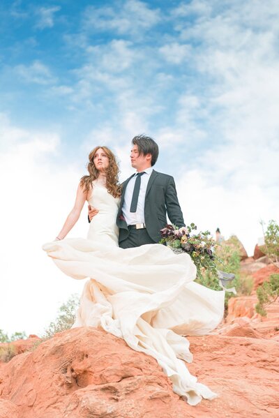 A bride and groom stand on a hill, looking off into the distance as the wind whips around them.