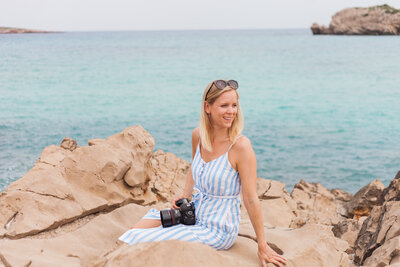 Philippa sitting on some rocks with her camera  by the sea in Menora after photographing a destination wedding