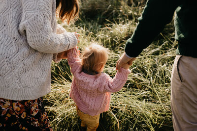A mother and father hold their child's hands and help her through a field