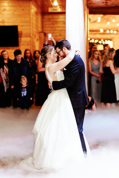 At a wedding captured by a Minnesota photographer, a couple dances closely. The bride in her white dress and the groom in his dark suit glide over the foggy floor. Guests in formal attire admire them, all set within a warmly lit, wood-paneled venue—a testament to exquisite Brainerd Lakes photography.