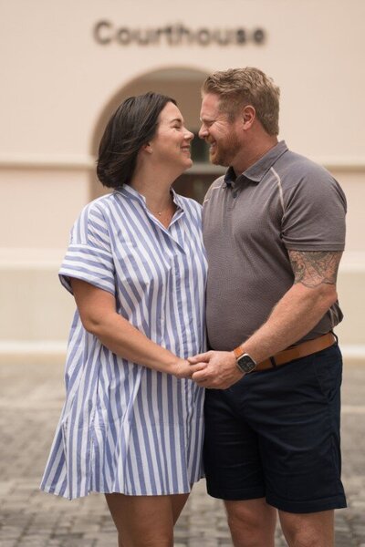 Happy couple embracing and smiling at each other in front of St. Augustine Courthouse during their wedding day. The woman wears a blue and white striped dress while the man wears a gray polo shirt and navy shorts.