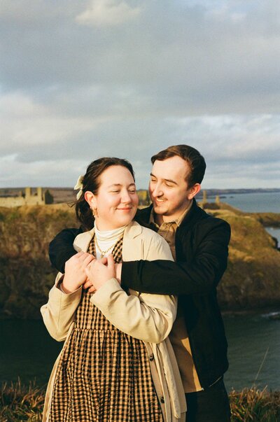 A couple hug each other back to front during their engagement photo session. An Aberdeenshire castle sits behind them on a cliff in the distance.