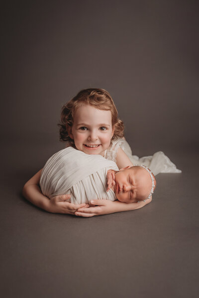 4 year old big sister in in white lace dress laying on her tummy smiling and holding newborn baby sister at newborn photo shoot at atlanta ga maternity and newborn photography studio
