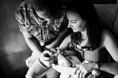 Parents sitting in and on the arm of a wooden rocking chair, mom holding son in crook of her arm and dad gently resting his hand on the baby's head