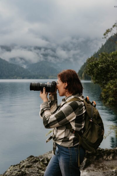 A person with a camera and backpack, like a Telluride Colorado wedding photographer, takes photos by a lake with mountains and cloudy skies in the background.