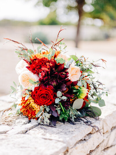 This image features a bouquet with red, burgundy, pink, and green hues propped up on a stone wall with a tree in the background. The image was taken by Austin wedding photographer KD Captures.