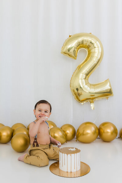 A little boy surrounded by gold balloons eats cake.
