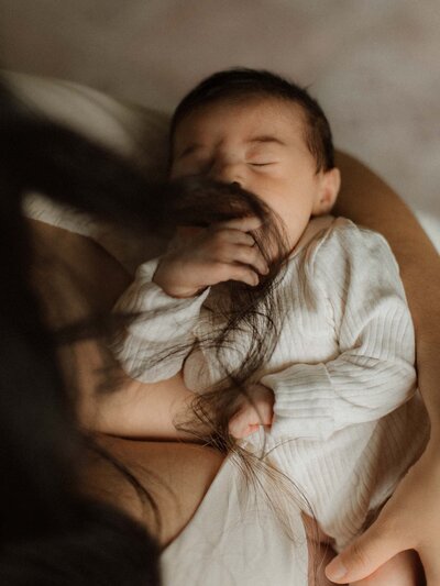 A newborn sleeps soundly with tiny fingers tangled in mama’s hair—an intimate in-home moment full of warmth and texture.