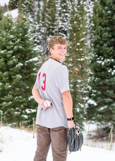 A high school senior boy wearing a gray baseball jersey holds a baseball and baseball glove while standing in front of snow covered pine trees at Tibble Fork Reservoir in American Fork