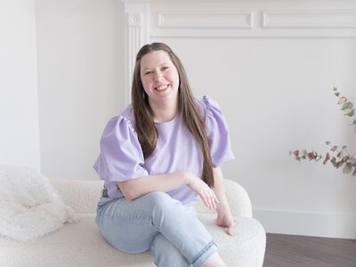 Emily Jane Lee wearing a purple blouse, sitting on a white boucle couch and smiling at camera.