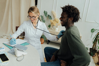 Healthcare provider in a white coat using a stethoscope on a smiling male patient who is standing.