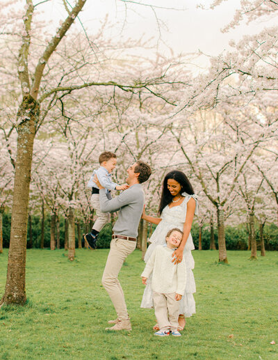 A dad throwing his younger son in the air, while mama and older son watch and laugh. 
