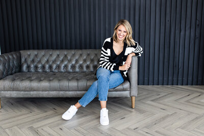 The woman is sitting on a dark gray sofa, and behind it is a textured black wall. She is dressed in a black and white striped textured jacket and a pair of jeans. Her arm is holding onto the sofa and she is laughing.