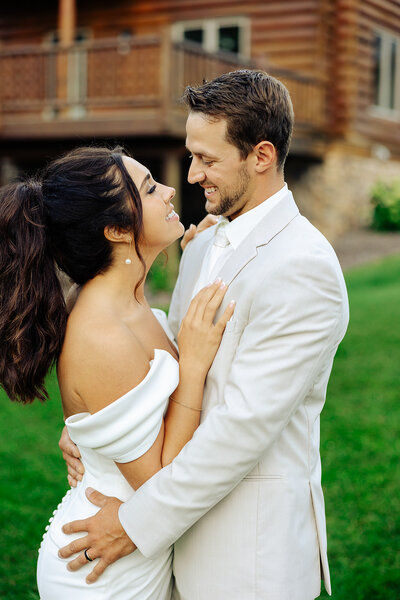 Captured by Amber Bauer Photography, a couple dressed in white smiles at each other outdoors. The woman wears an off-shoulder dress, and the man wears a suit as they stand on a grassy area with a wooden building in the background.