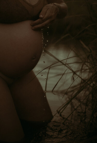 A pregnant woman is up to her knees in water. You see her pregnant belly in the middle of the frame with water dripping down from her hand