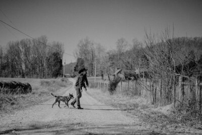 Cowgirl walks across gravel road with her cattle dog trotting behind her..