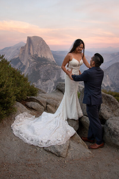 Newlywebs embrace in the setting sunlight with Yosemite's Half Dome standing tall in the background.