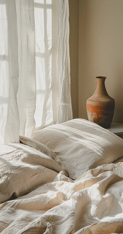 Gauzy white curtains in front of window in front of the bed covered in flaxen colored linens with medium sized pottery jar on side table