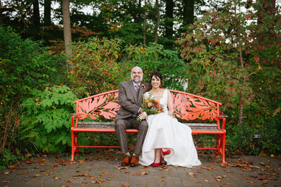 couple sitting on a red bench in a park on their wedding day